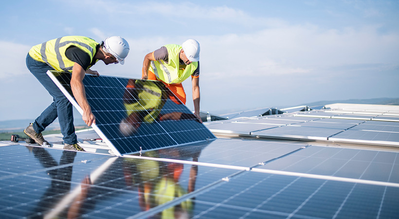 Two workers wearing white hard hats and bright yellow vests install solar panels on a rooftop.