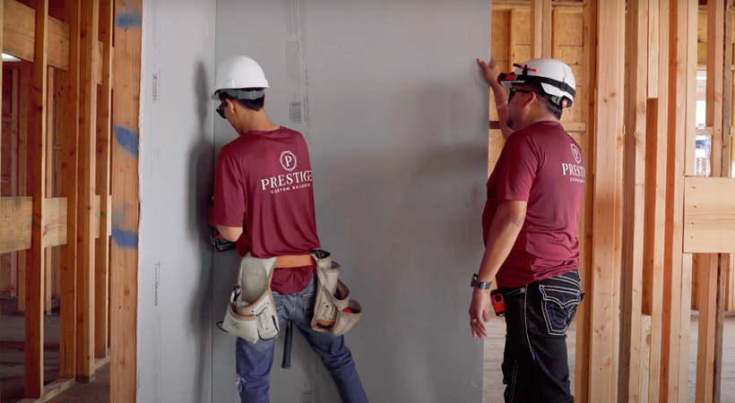 Two construction workers, wearing white hard hats and maroon Prestige Custom Builders shirts, install DensShield® Tile Backer on framing.