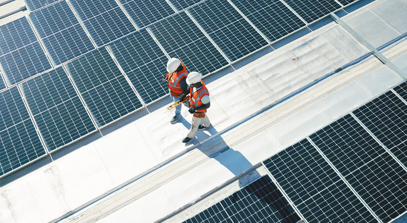 An aerial view of 2 workers wearing white hard hats and bright orange vests walking and talking between solar panels on a rooftop. 