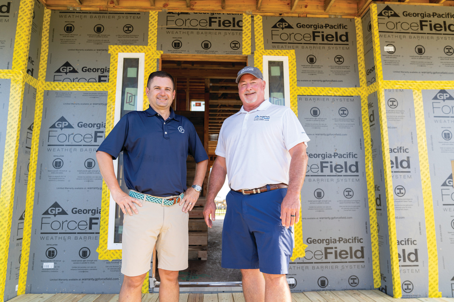 Two men in shorts and polo shirts stand in front of a home sheathed in ForceField® Weather Barrier System. They are smiling.