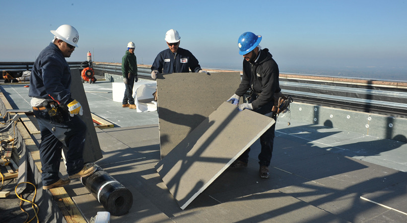 Several roofers wearing hard hats, boots and construction gear install cover boards on a rooftop.